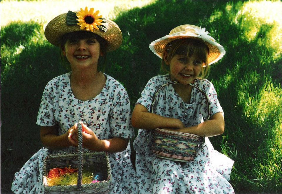A picture of 2 girls sitting on the grass, one brunette and one blonde, wearing matching dresses and hats, smiling and holding easter baskets.